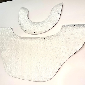May include: Two white, textured, semi-circular leather pieces with a distinct ostrich-like pattern. The larger piece is wide and curved, while the smaller piece is a narrow crescent. Both items feature evenly spaced small holes along their edges.