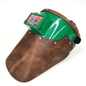 May include: Green and brown welding helmet with a textured leather-like face shield. The helmet has a rectangular display with colorful graphics and the text "KEEPER CLASSY". Rivets secure the face shield to the green helmet.