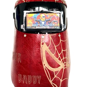 May include: A red and black welding helmet with a gold spiderweb design and the words "YOUR DADDY". The helmet features a screen with the text "KEEP'ER CLASSY" and a cartoon image of a welder.