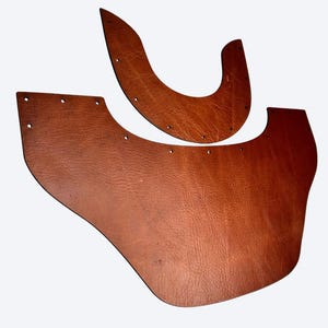 May include: Two pieces of brown leather with a textured surface and visible grain. The larger piece is wide with a curved bottom edge and multiple pre-punched holes along the top. The smaller piece is crescent-shaped with holes along the edge.