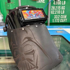 May include: A black welding helmet with a Darth Vader design etched on the front. The helmet has a rectangular viewing screen with the text "KEEP'ER CLASSY" and a colorful graphic. The helmet is designed for welding.