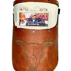 May include: Welding helmet with a brown leather exterior and a white top. The helmet features a bull skull design and the text "KEEP'ER CLASSY" on the viewing screen. The helmet is designed for welding and offers protection for the eyes.