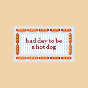 May include: A rectangular graphic print featuring a light blue background with vertical white stripes. The text "bad day to be a hot dog" is centered in red pixelated font. A border of small, repeating hot dog icons surrounds the text.