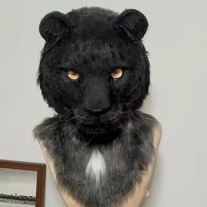 May include: A black panther mask with realistic fur details. The mask features golden eyes, a detailed nose, and a faux fur mane in shades of gray and white. The mask is designed to be worn and is displayed on a mannequin head.