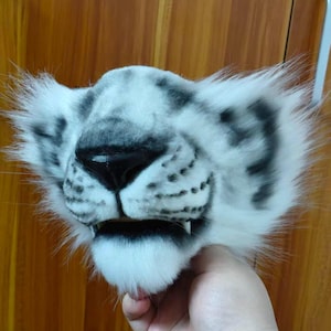 May include: A white and gray snow leopard mask with black spots and a black nose. The mask has fluffy white fur around the ears and mouth, and small teeth are visible. The mask is held by a hand.