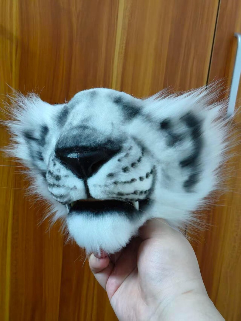 May include: A white and gray snow leopard mask with black spots and a black nose. The mask has fluffy white fur around the ears and mouth, and small teeth are visible. The mask is held by a hand.