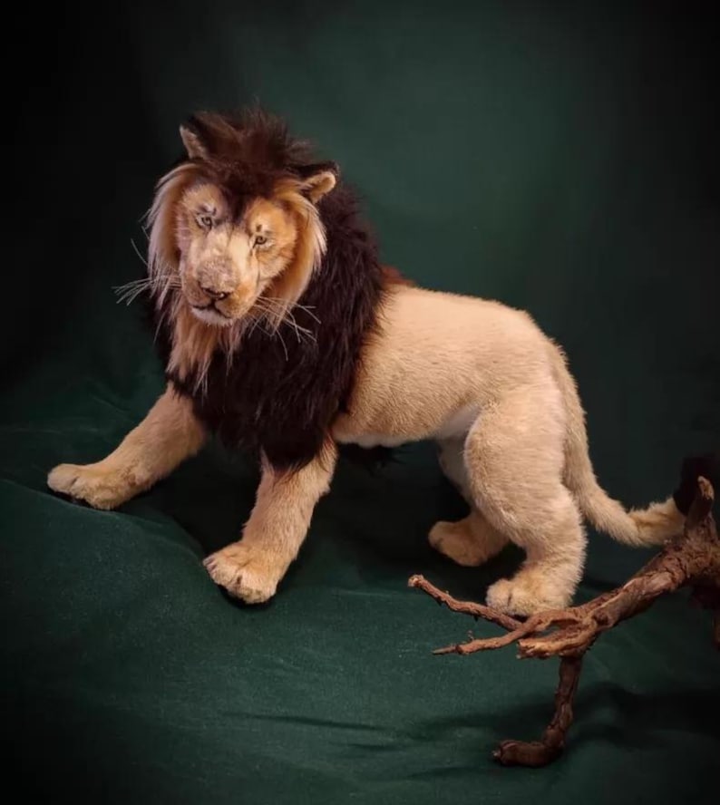 Pode incluir: Um le&atilde;o de pel&uacute;cia realista com corpo bege e juba castanha escura. O le&atilde;o est&aacute; posicionado sobre uma superf&iacute;cie verde, com rosto detalhado e textura de pelo realista. Um pequeno peda&ccedil;o de madeira est&aacute; em primeiro plano.