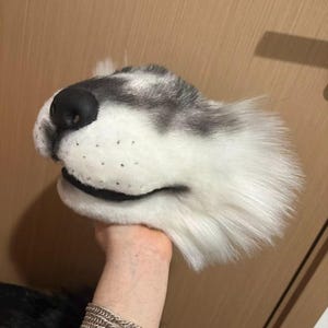 Op de afbeelding: Een pluche hondenhoofd met een zwarte neus en grijs en witte vacht. De bek van de hond staat open en de vacht is zacht en pluizig. Het hoofd wordt vastgehouden door een hand, met een houten deur op de achtergrond.