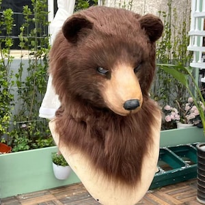 Può includere: Una maschera realistica a testa di orso bruno con folta pelliccia sintetica testurizzata, un muso beige modellato, un naso nero e occhi stretti e concentrati. La maschera è montata su un supporto, mettendo in risalto la texture del pelo.