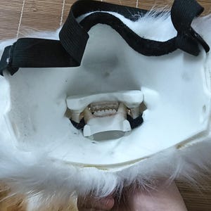 May include: A white and orange furry mask with a black elastic strap. The mask has a detailed mouth with teeth and is designed to be worn on the face. The mask is likely for cosplay or costume use.