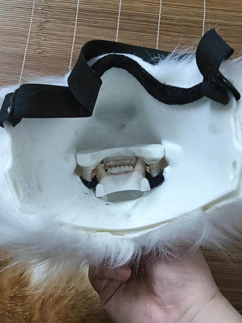 May include: A white and orange furry mask with a black elastic strap. The mask has a detailed mouth with teeth and is designed to be worn on the face. The mask is likely for cosplay or costume use.