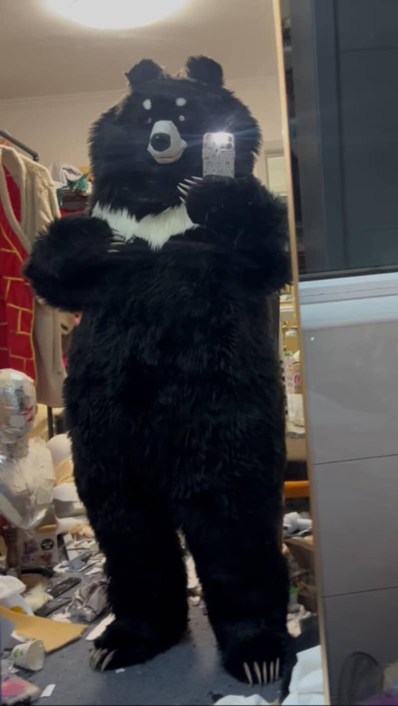 May include: A full-body black bear costume with a white chest patch. The bear suit has a plush, furry texture, with a detailed head featuring a snout, eyes, and ears. The person wearing the costume is holding a phone.
