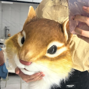 May include: A plush chipmunk mask with brown, tan, and white fur. The mask has large black eyes, a pink nose, and long whiskers. The mask is being held up by a person.