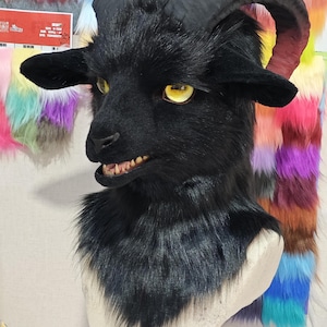 May include: A black goat mask with large curved horns and yellow eyes. The mask has a furry texture and detailed facial features, including visible teeth. The background features colorful fabric swatches.