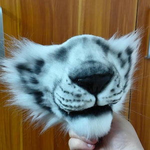 May include: A white and gray tiger mask with black markings around the nose and mouth. The mask has fluffy white fur around the edges and is held up by a hand.