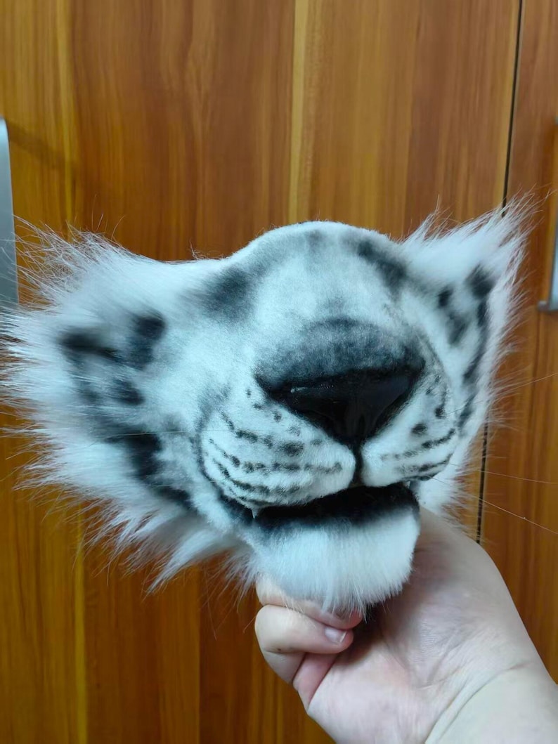 May include: A white and gray tiger mask with black markings around the nose and mouth. The mask has fluffy white fur around the edges and is held up by a hand.