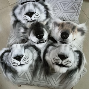 May include: Five plush lion masks with black noses, white fur, and gray accents are arranged on a chair. The masks have open mouths and are designed to be worn. The chair has a gray geometric pattern.