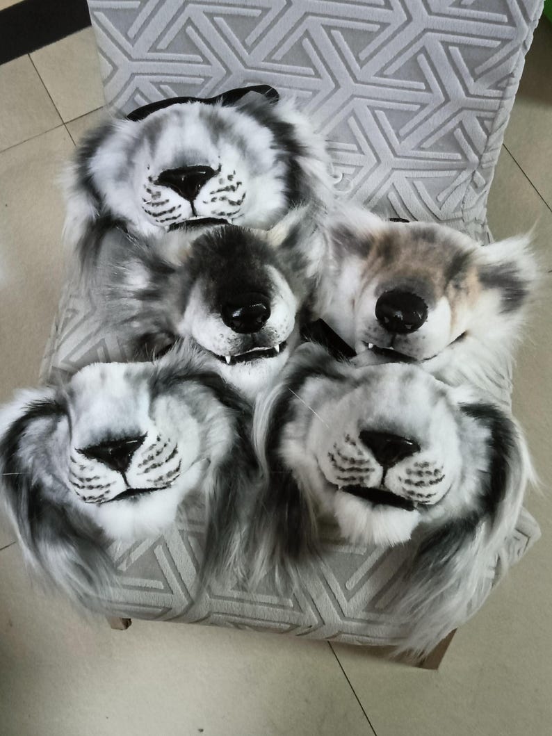 May include: Five plush lion masks with black noses, white fur, and gray accents are arranged on a chair. The masks have open mouths and are designed to be worn. The chair has a gray geometric pattern.