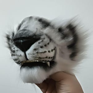 May include: A white and gray snow leopard mask with black accents. The mask features a realistic design with detailed fur texture, a black nose, and visible teeth. The ears are fluffy and the overall design is lifelike.