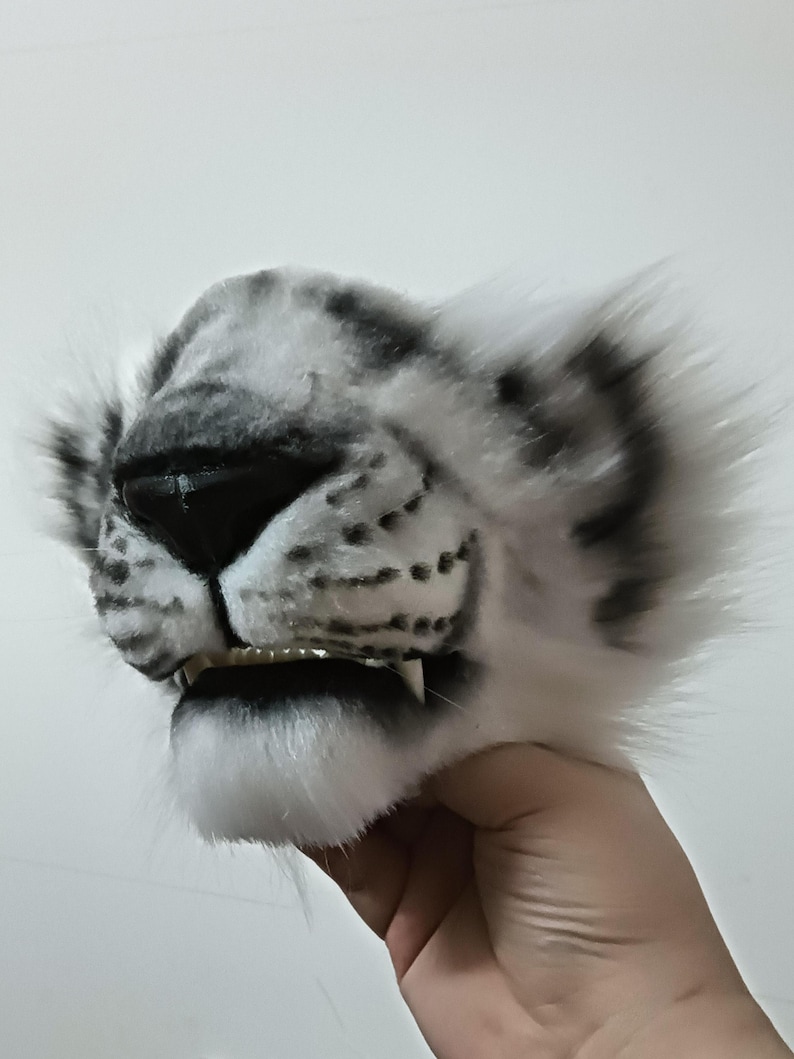 May include: A white and gray snow leopard mask with black accents. The mask features a realistic design with detailed fur texture, a black nose, and visible teeth. The ears are fluffy and the overall design is lifelike.