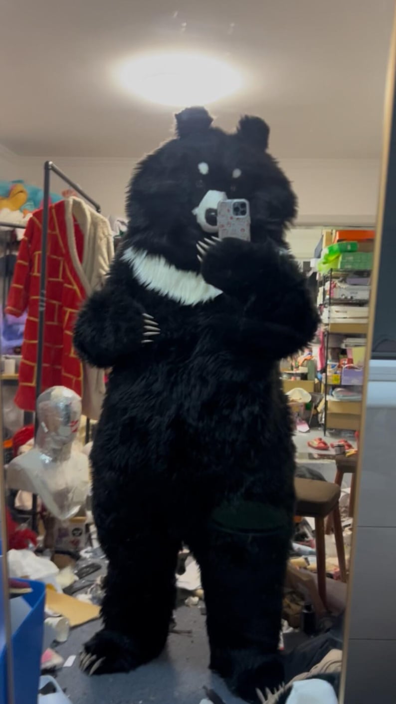 May include: A full-body black bear costume with a white chest marking. The furry costume features large paws with claws, a bear head with ears, and a white muzzle. The person wearing the costume is holding a phone in front of their face.