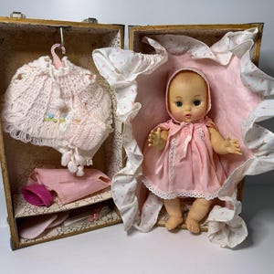 May include: A vintage doll in a pink dress and bonnet, presented in an open decorative case. The case contains the doll and accessories, such as a pink knitted cape and other items. The doll has a sweet expression.