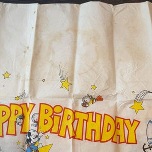 May include: Vintage birthday banner with a cream background and yellow and red text that reads "HAPPY BIRTHDAY". The banner features cartoon characters, stars, and shooting stars. The banner is folded and has some stains.