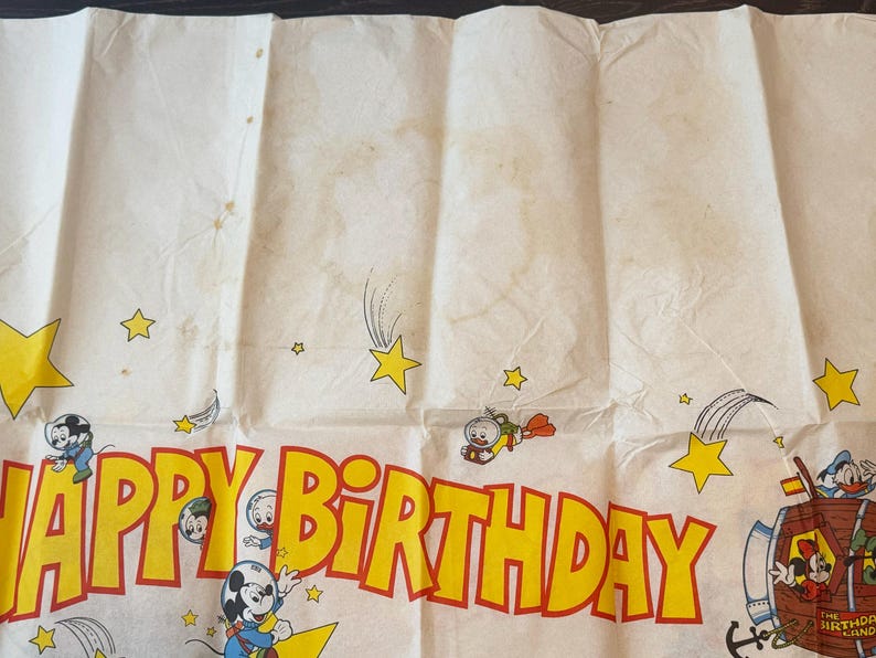 May include: Vintage birthday banner with a cream background and yellow and red text that reads "HAPPY BIRTHDAY". The banner features cartoon characters, stars, and shooting stars. The banner is folded and has some stains.