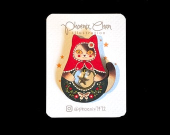 Può includere: Spilla in legno con un gatto che indossa un mantello rosso con cappuccio e tiene una stella dorata. Il gatto è circondato da dettagli floreali e farfalle. La spilla è su una carta bianca con il testo "Phoenix Chan Illustration" e "@phoenix7472".