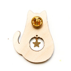 May include: Wooden cat pin with a gold star charm hanging from the cat's belly. The pin has a gold back with a safety clasp.