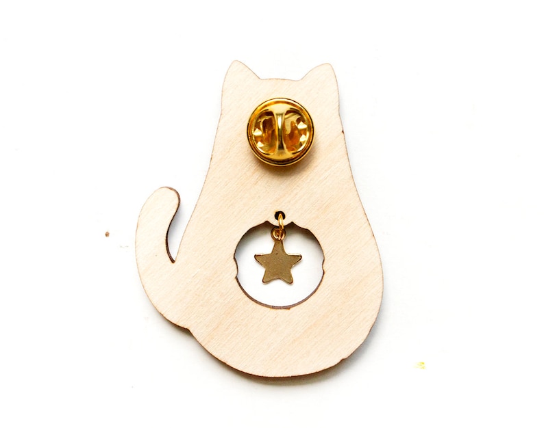 May include: Wooden cat pin with a gold star charm hanging from the cat's belly. The pin has a gold back with a safety clasp.