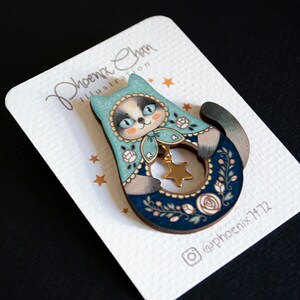 May include: A wooden pin featuring a blue and white cat holding a gold star. The cat is wearing a blue and white hat with white flowers. The pin is on a white card with the text "Phoenix Chen Illustration" and "@phoenix7172".