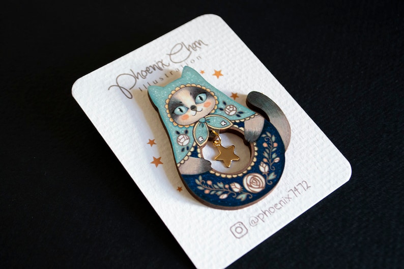 May include: A wooden pin featuring a blue and white cat holding a gold star. The cat is wearing a blue and white hat with white flowers. The pin is on a white card with the text "Phoenix Chen Illustration" and "@phoenix7172".