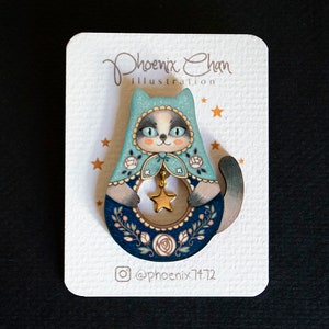 May include: A wooden cat-shaped pin with a blue and white hooded design. The cat is holding a gold star and has a floral pattern around the bottom. The pin is on a white card with the text "Phoenix Chan Illustration" and "@phoenix7172".