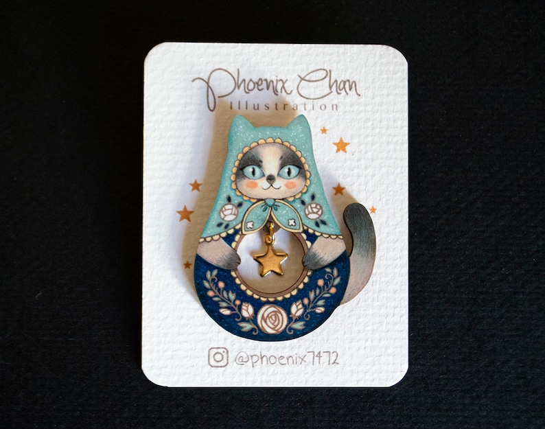 May include: A wooden cat-shaped pin with a blue and white hooded design. The cat is holding a gold star and has a floral pattern around the bottom. The pin is on a white card with the text "Phoenix Chan Illustration" and "@phoenix7172".
