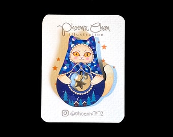 Può includere: Uno spillo di legno a forma di gatto con un design a motivi blu e bianchi. Il gatto indossa un cappello blu e bianco e tiene una stella dorata. Lo spillo è su una carta bianca con il testo "Phoenix Chan Illustration" e "@phoenix_7472".