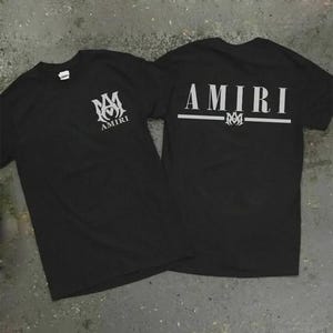 Puede incluir: Dos camisetas negras con la palabra "AMIRI" impresa en blanco. Una camiseta tiene el logotipo y el texto en la parte delantera, y la otra tiene el texto y el logotipo en la parte trasera. Las camisetas est&aacute;n hechas de un material suave.