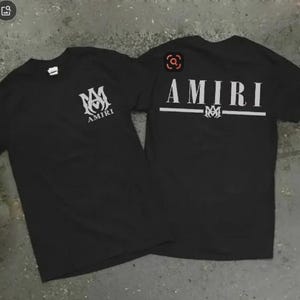 Puede incluir: Dos camisetas negras con la palabra "AMIRI" impresa en blanco. Una camiseta tiene el logo en la parte delantera y la otra en la trasera. Las camisetas son de manga corta y parecen estar hechas de un material suave.