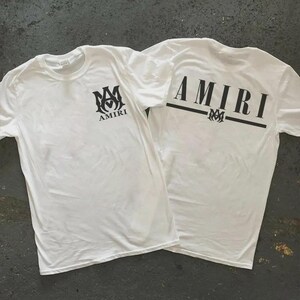Puede incluir: Dos camisetas blancas con logotipos AMIRI negros. Una camiseta tiene el logotipo en la parte delantera y la otra tiene la palabra "AMIRI" en la espalda. Las camisetas est&aacute;n hechas de un material suave y ligero.