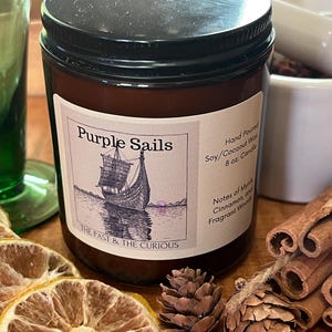 May include: A brown glass jar candle with a black lid and a label that reads "Purple Sails." The label features a drawing of a ship. The candle is 227g and made with soy/coconut wax. Cinnamon sticks and dried lemon slices are also visible.