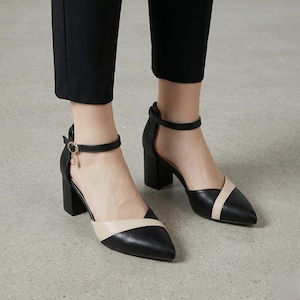 Könnte beinhalten: Schwarze und beige spitz zulaufende Pumps mit Knöchelriemen und Blockabsätzen. Die Schuhe haben einen kontrastierenden beigefarbenen Einsatz über dem Spann und einen kleinen goldenen Anhänger am Knöchelriemen. Die Absätze sind ca. 7,6 cm hoch.