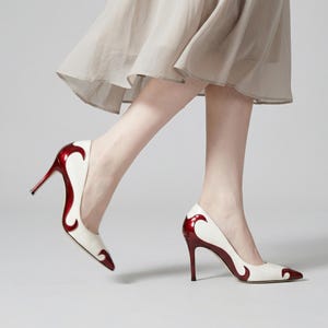 May include: White and red high-heeled pumps. The shoes have a pointed toe and a slender heel. The red design curves along the side of the shoe. The pumps are worn with a beige skirt.