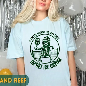 Puede incluir: Una camiseta azul claro con un gráfico verde oscuro de un personaje de pepinillo enojado sosteniendo una pala de pickleball frente a una red. El texto dice: IF YOU ARE LOOKING FOR SOFT SERVE GO GET ICE CREAM. El diseño es circular y centrado.