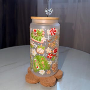 Könnte beinhalten: Ein mattierter Glastrinkbecher mit Bambusdeckel und einem klaren Glasstrohhalm mit einer dekorativen Schleife. Die Außenseite ist mit einem festlichen Muster aus grünen Pullovern, Lebkuchen, Zuckerstangen, Geschenken und Weihnachtssternen bedruckt.