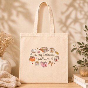 Könnte beinhalten: Eine cremefarbene Stofftasche mit einem zentralen Aufdruck, der den Text 'in my bookish fall era' zeigt, umgeben von Illustrationen von Büchern, einem Kürbis, einer Kerze, einer Tasse, einer Zimtschnecke und einer Schleife in sanften Farben.