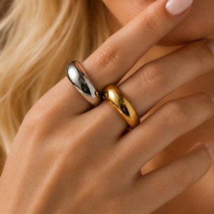 May include: Two smooth, thick, rounded bands worn on adjacent fingers. One ring features a polished silver-tone finish, while the other ring displays a high-shine gold-tone finish. Both pieces have a simple, solid, and continuous circular design.