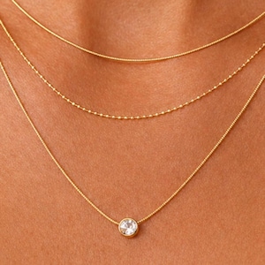 May include: A layered gold necklace set consisting of three separate chains. The top chain is a smooth gold cable, the middle chain features small gold beads, and the bottom chain holds a single round clear stone in a gold bezel setting.