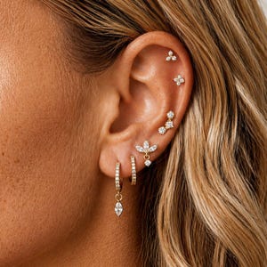May include: A set of gold-toned earrings featuring multiple small hoops and studs with clear stones. The jewelry includes a dangling marquise-cut stone, a floral-shaped stud, and several small circular studs arranged along the ear cartilage.