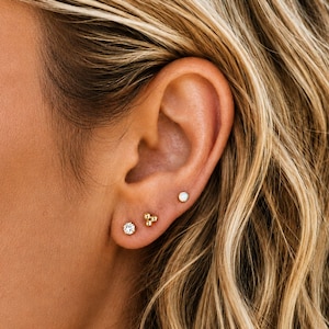 May include: Three small stud earrings worn in a single earlobe. The top earring is a simple white sphere, the middle earring features three tiny gold-toned beads arranged in a triangle, and the bottom earring is a single clear round gemstone.