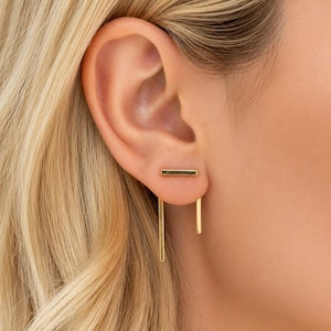 May include: A gold-toned ear jacket earring featuring a horizontal bar stud with a dark green inlay. Two thin, vertical gold-toned bars of unequal length extend downwards from the back of the earlobe, creating a modern, geometric, and layered appearance.
